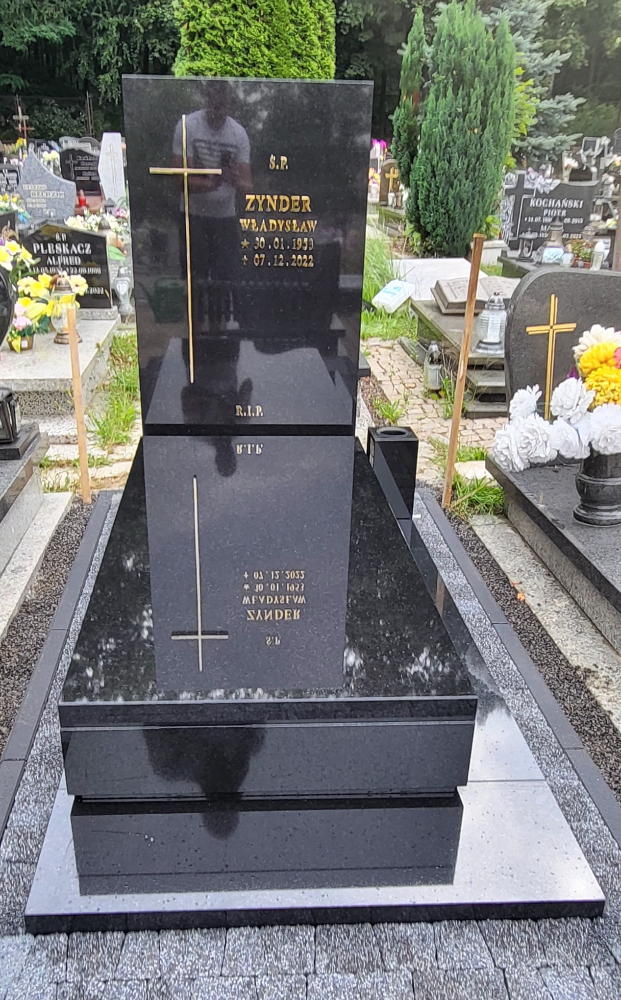 Czarny, polerowany pomnik z wygrawerowanym złotym krzyżem i inskrypcjami, otoczony innymi nagrobkami na cmentarzu.