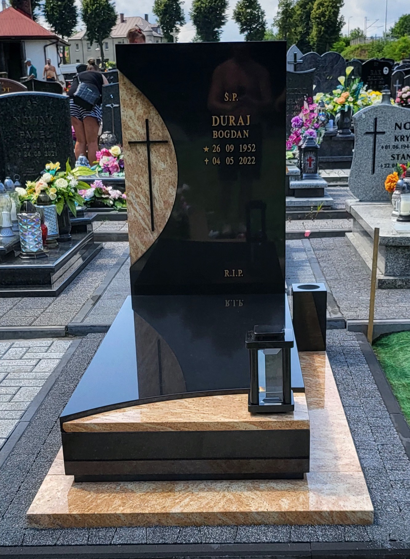 Nowoczesny, czarny pomnik z elementami beżowego kamienia i krzyżem, na cmentarzu; widoczna zapalona lampion i inne nagrobki w tle.