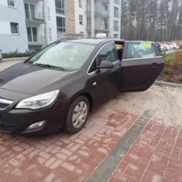 Brązowy samochód osobowy z otwartymi drzwiami, oznaczony jako taksówka, zaparkowany na kostce brukowej przed nowoczesnym budynkiem mieszkalnym.