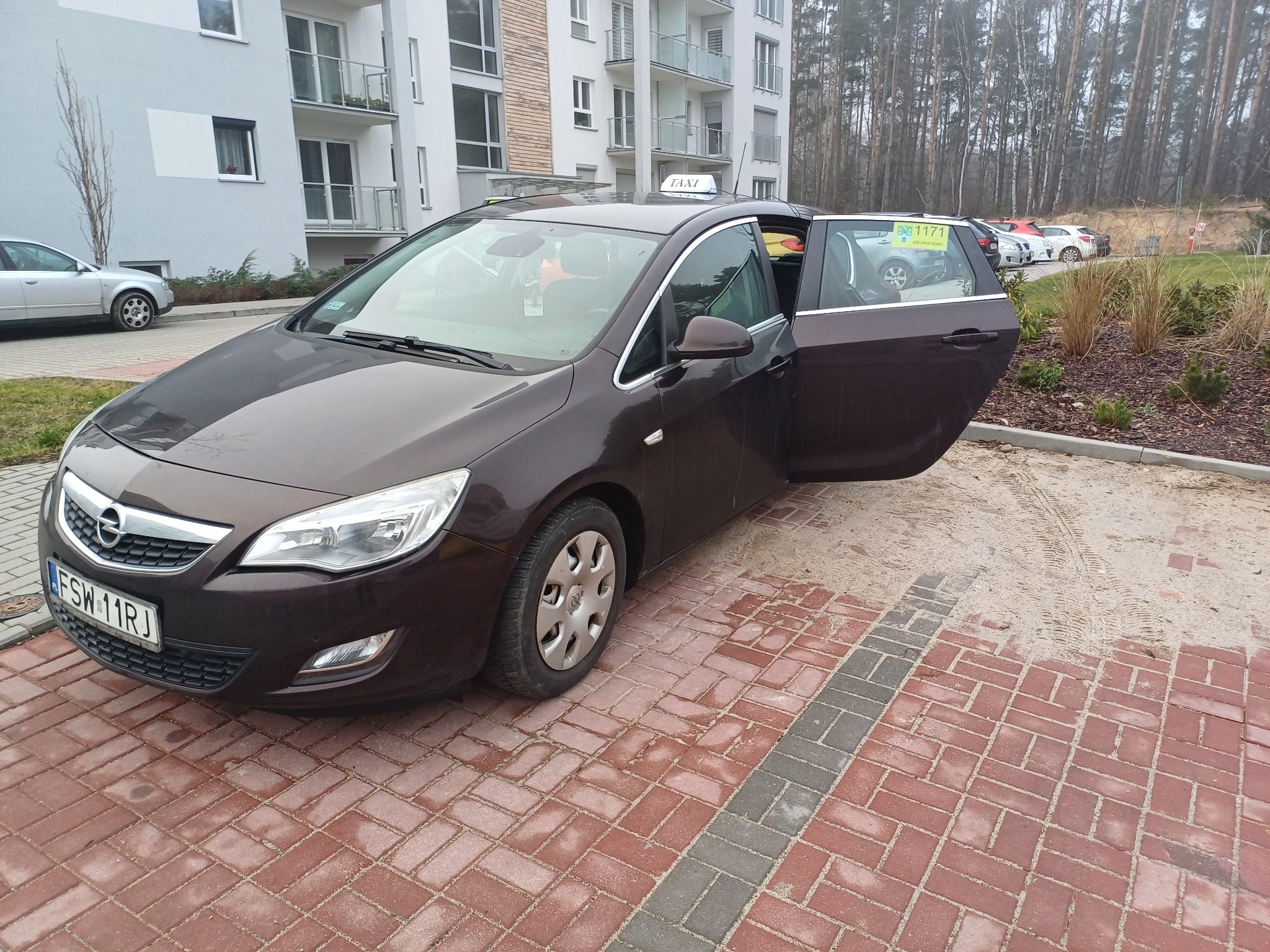 Brązowy samochód osobowy z otwartymi drzwiami, oznaczony jako taksówka, zaparkowany na kostce brukowej przed nowoczesnym budynkiem mieszkalnym.