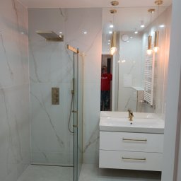 Budex - Nowoczesna łazienka z kabiną prysznicową typu walk-in, wykończona białymi płytkami imitującymi marmur z delikatnymi złotymi akcentami w armaturze i wykończeniach, widoczna umywalka z białą szafką...