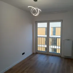 Pusty pokój z drewnianą podłogą, białymi ścianami, nowoczesną spiralną lampą sufitową i oknem balkonowym z widokiem na budynek.