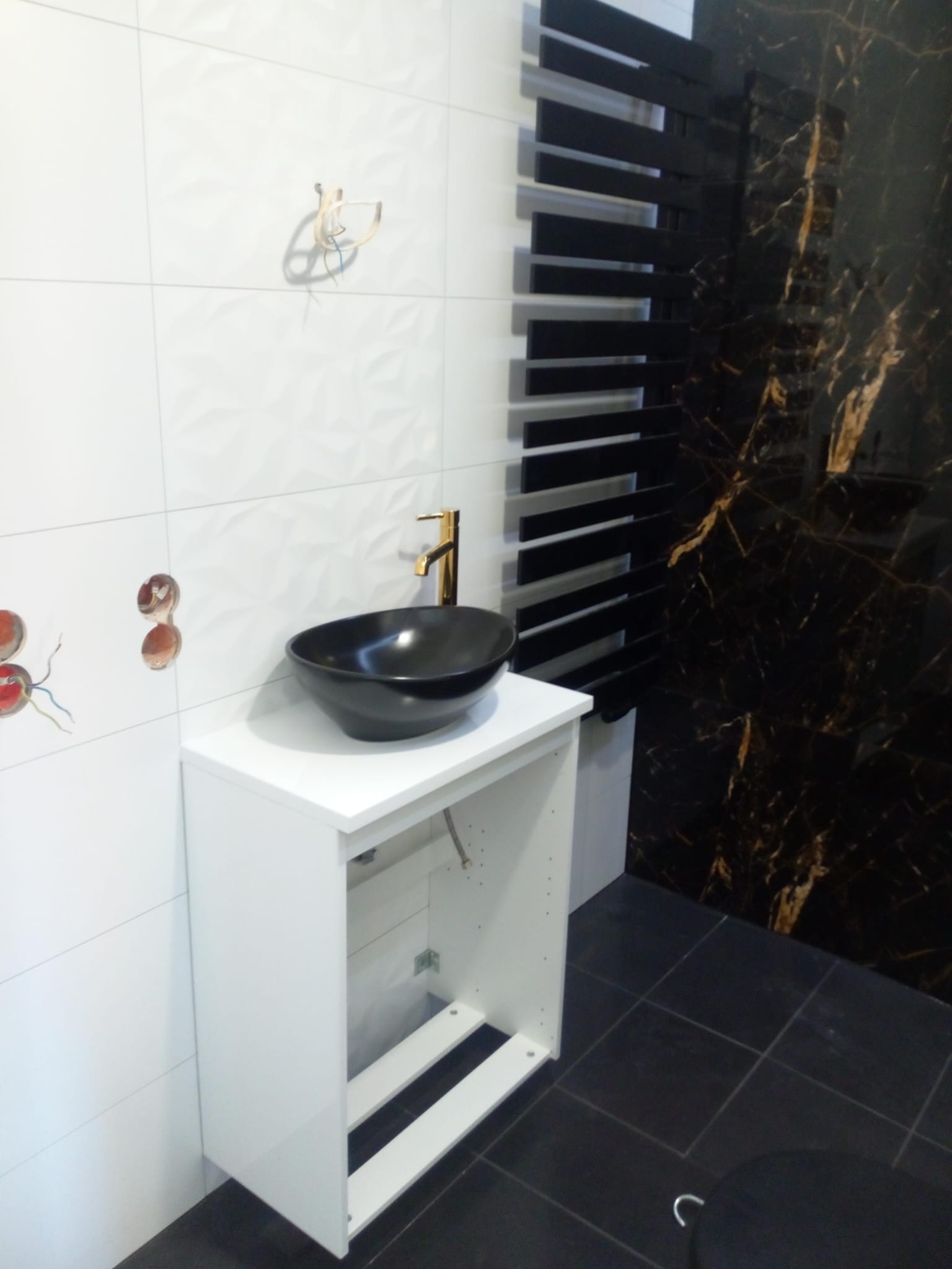 Czarna, okrągła umywalka nablatowa z złotą baterią na białej szafce łazienkowej. W tle białe płytki z geometrycznym wzorem, czarny grzejnik drabinkowy i płytki imitujące czarny marmur ze złotymi...