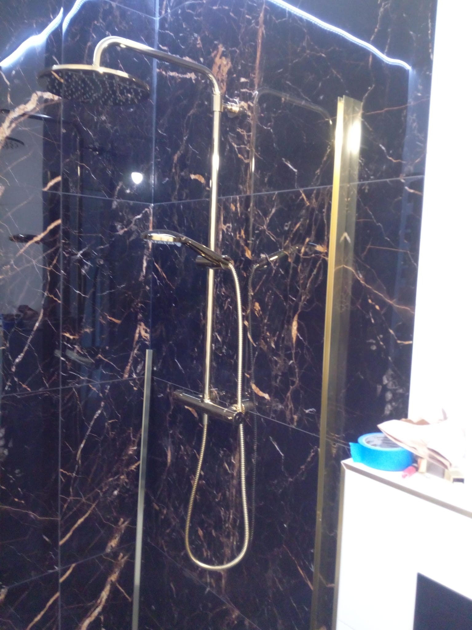 Nowoczesna kabina prysznicowa z deszczownicą, wykończona czarnymi płytkami imitującymi marmur ze złotymi żyłami, z widocznymi elementami montażowymi i fragmentem szafki z niebieską taśmą malarską.