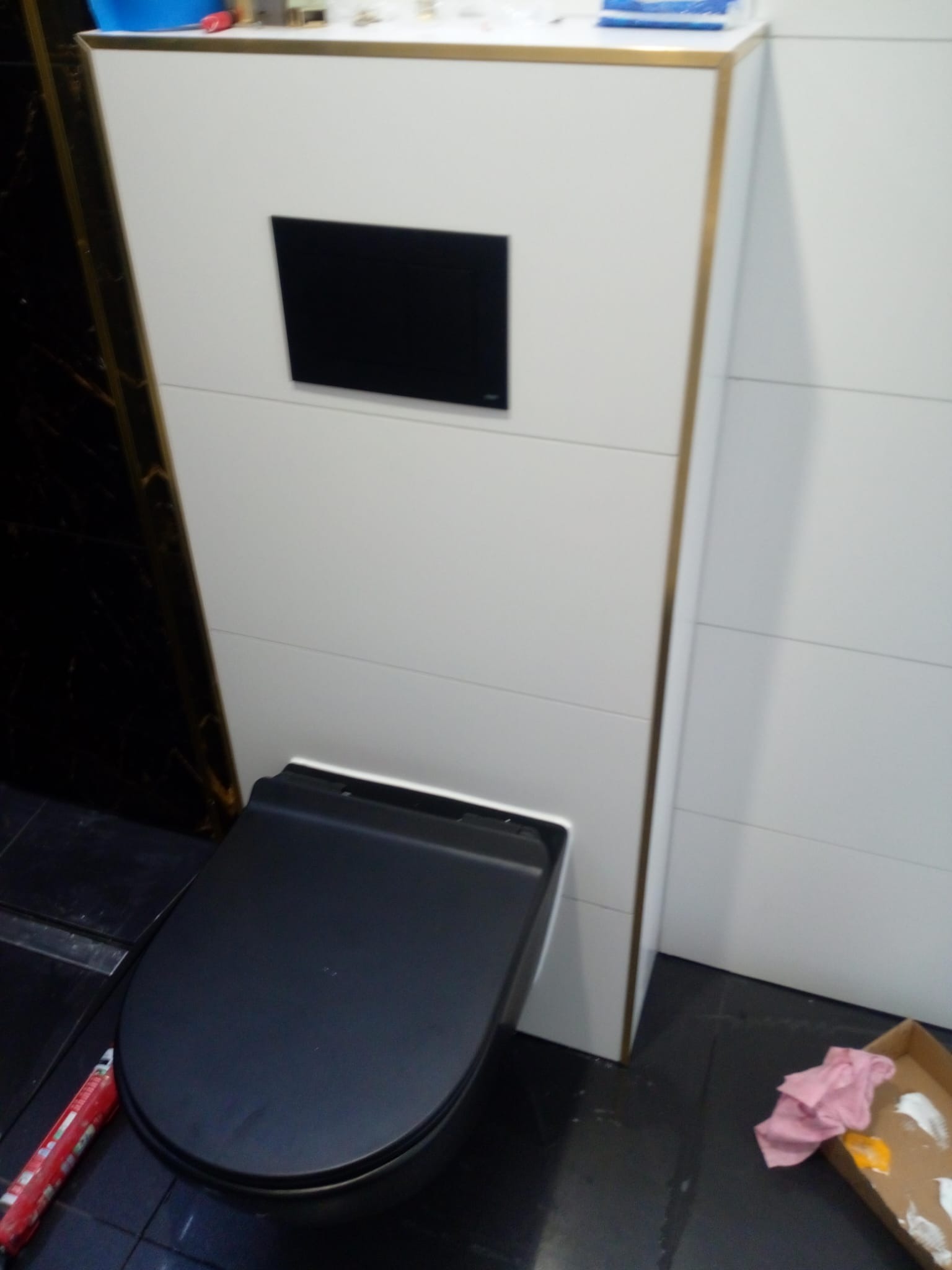 Czarna, wisząca toaleta z ukrytym zbiornikiem w zabudowie z białych płyt z ozdobnymi złotymi listwami, czarne płytki podłogowe, widoczne narzędzia i akcesoria malarskie.