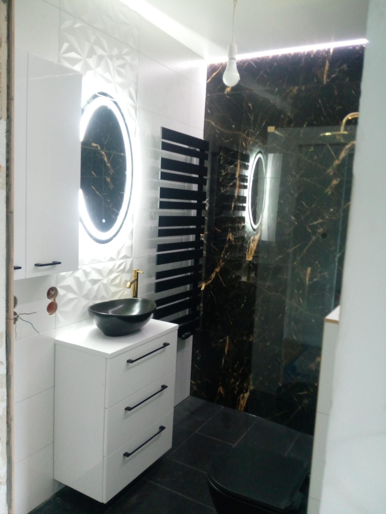 Nowoczesna łazienka z czarną umywalką na białej szafce z trzema szufladami, okrągłe lustro z podświetleniem LED, grzejnik drabinkowy, czarne płytki imitujące marmur z złotymi żyłami w kabinie...