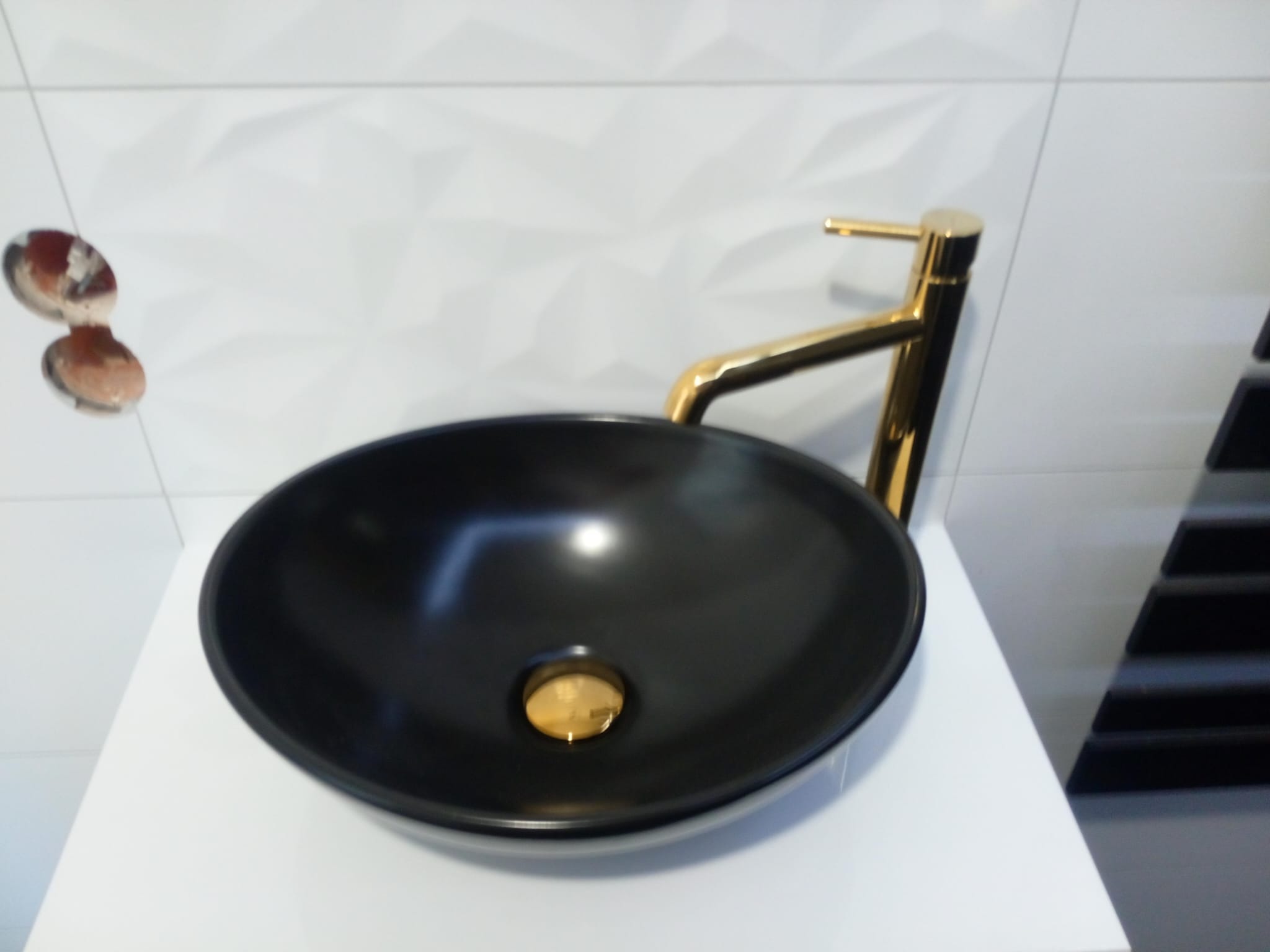 Czarna, okrągła umywalka nablatowa z złotą baterią na białym blacie w łazience w Węglińcu, z widocznymi niedokończonymi otworami w ścianie za umywalką.