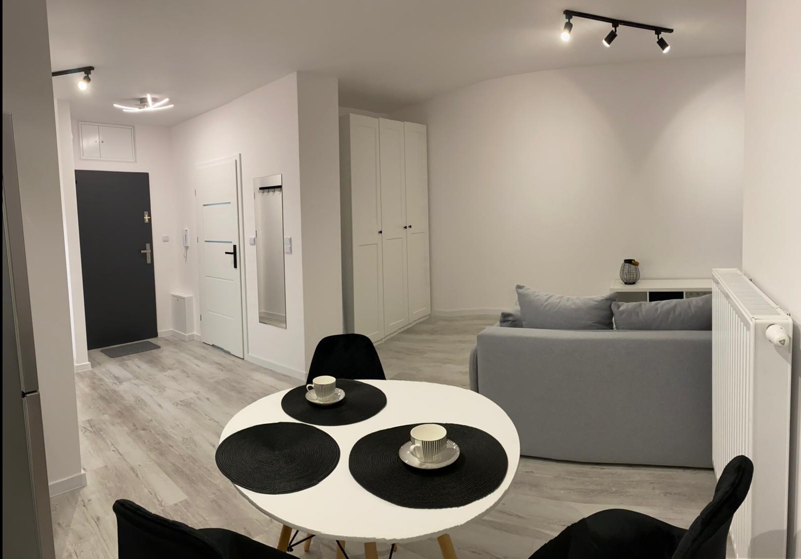 Jasne wnętrze pokoju po remoncie z białymi ścianami, panelami podłogowymi, szarą sofą, białym stołem z czarnymi podkładkami i filiżankami oraz białymi szafami w zabudowie.