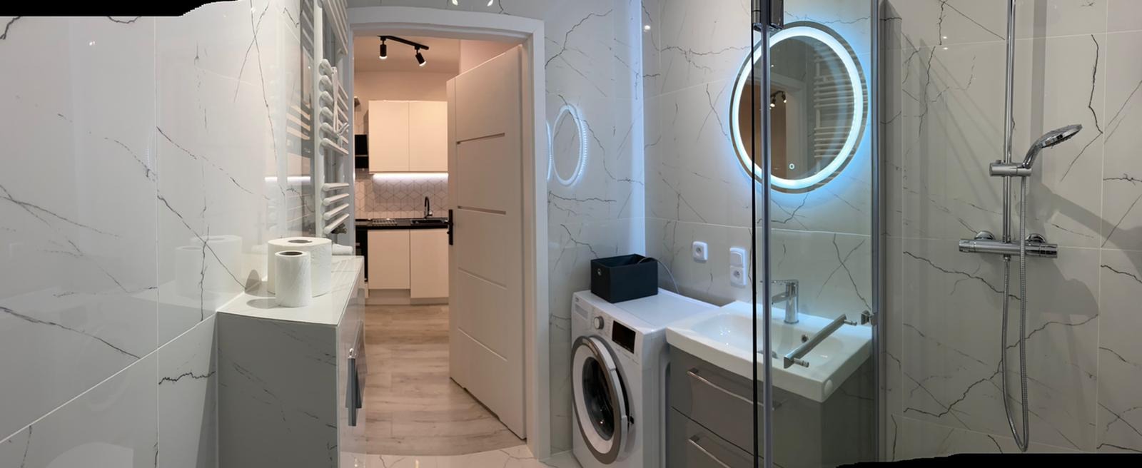 Nowoczesna łazienka z białymi płytkami imitującymi marmur, okrągłym lustrem z podświetleniem LED, kabiną prysznicową ze szklaną ścianką, pralką pod umywalką i widokiem na aneks kuchenny przez...