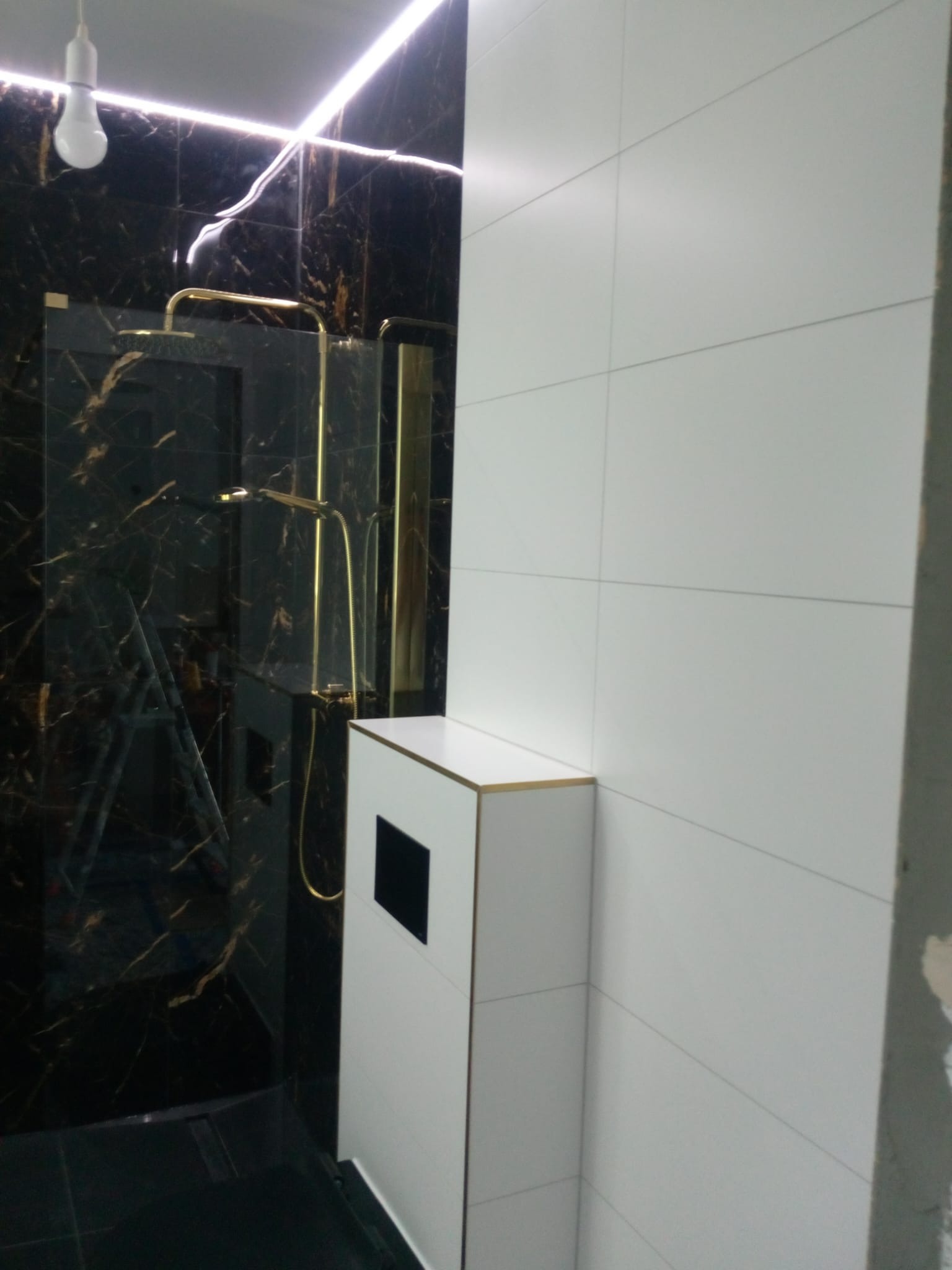 Nowoczesna łazienka z czarnymi marmurowymi płytkami i złotą armaturą prysznicową, biała zabudowa podtynkowa WC z czarnym panelem, oświetlenie LED pod sufitem, drabina w tle.