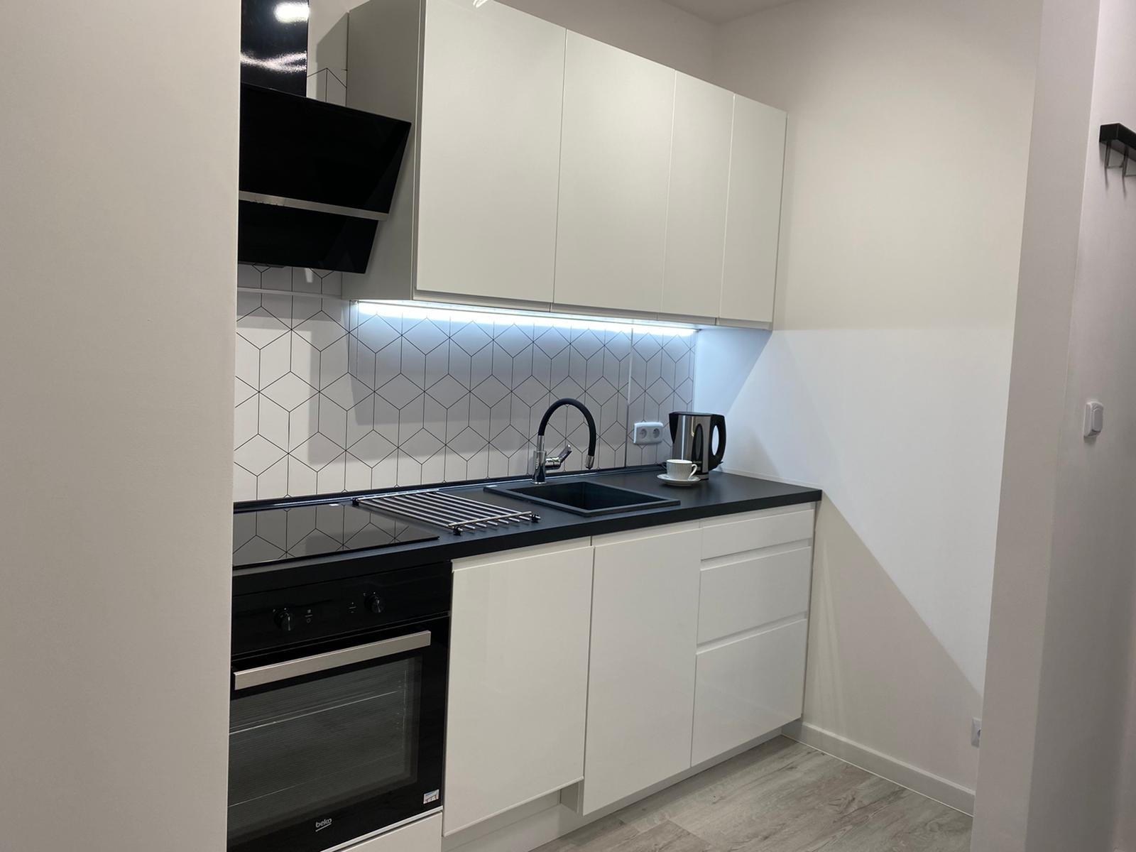 Nowoczesna kuchnia z białymi szafkami, czarnym blatem i geometrycznymi płytkami na ścianie, oświetlona listwą LED pod szafkami wiszącymi, z widocznym piekarnikiem Beko.