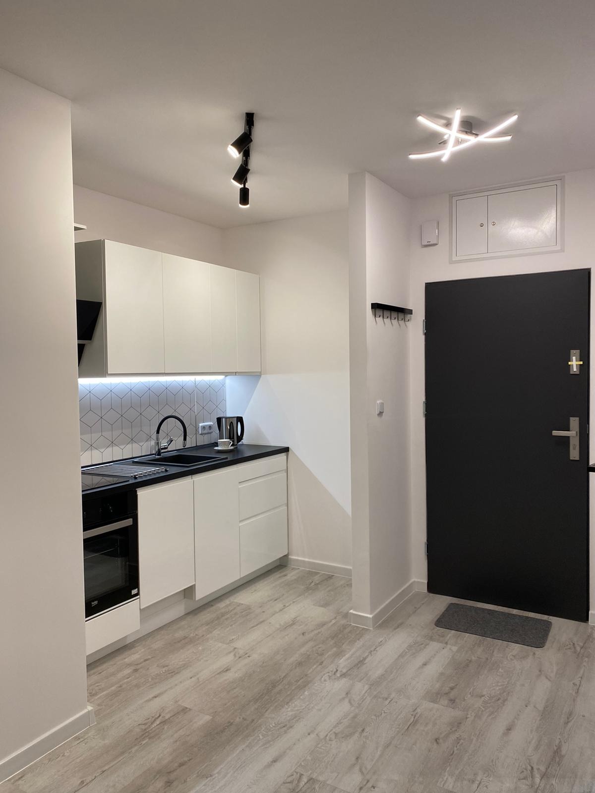 Nowoczesna, minimalistyczna kuchnia w odcieniach bieli i czerni, z drewnopodobną podłogą, oświetleniem punktowym i geometryczną mozaiką nad blatem, widziana z perspektywy wejścia do mieszkania.