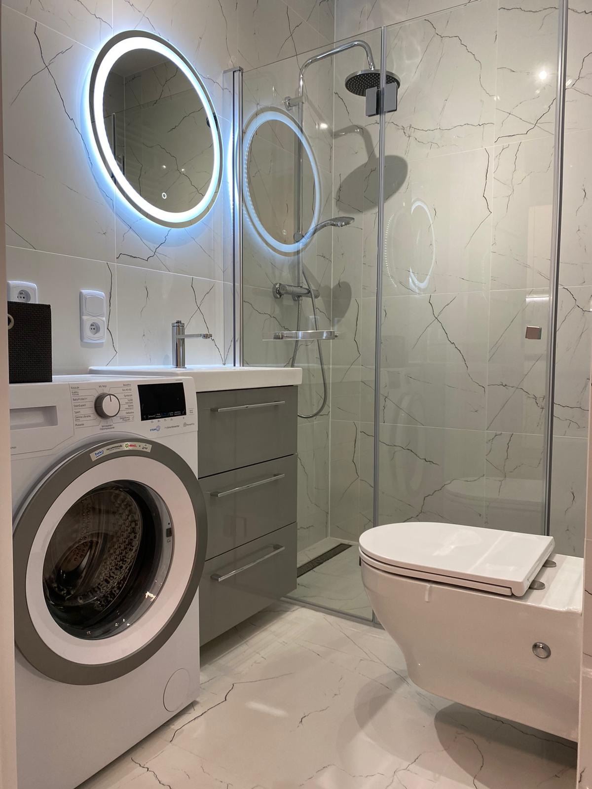 Nowoczesna łazienka z kabiną prysznicową, toaletą wiszącą, umywalką z szufladami i pralką, wykończona płytkami imitującymi marmur, z okrągłymi lustrami podświetlanymi LED.