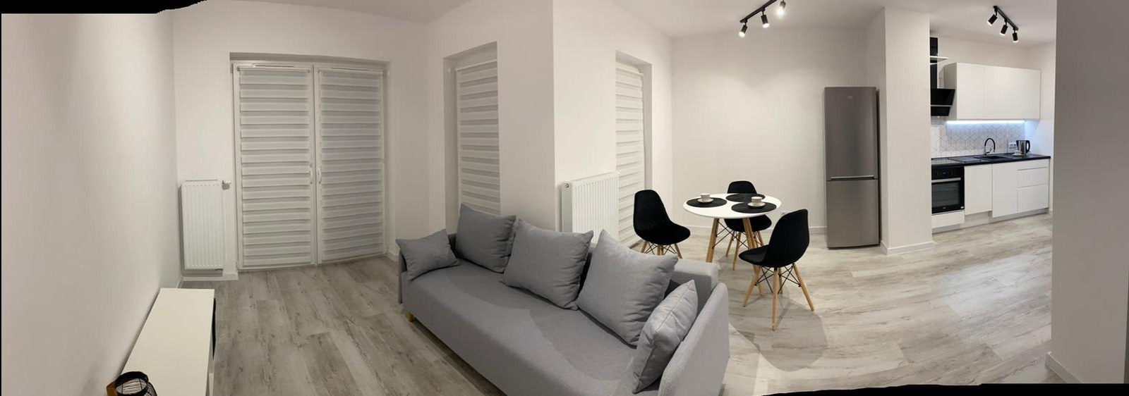 Panoramiczny widok nowocześnie urządzonego mieszkania: szara sofa, okrągły stół z czarnymi krzesłami, biała kuchnia z czarnymi akcentami, jasne ściany i podłoga imitująca drewno.