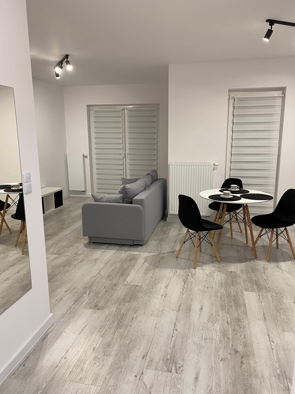 Jasne, minimalistyczne wnętrze pokoju dziennego z szarą sofą, okrągłym białym stolikiem i czarnymi krzesłami na drewnianych nogach, podłoga wyłożona panelami imitującymi jasne drewno, białe ściany...