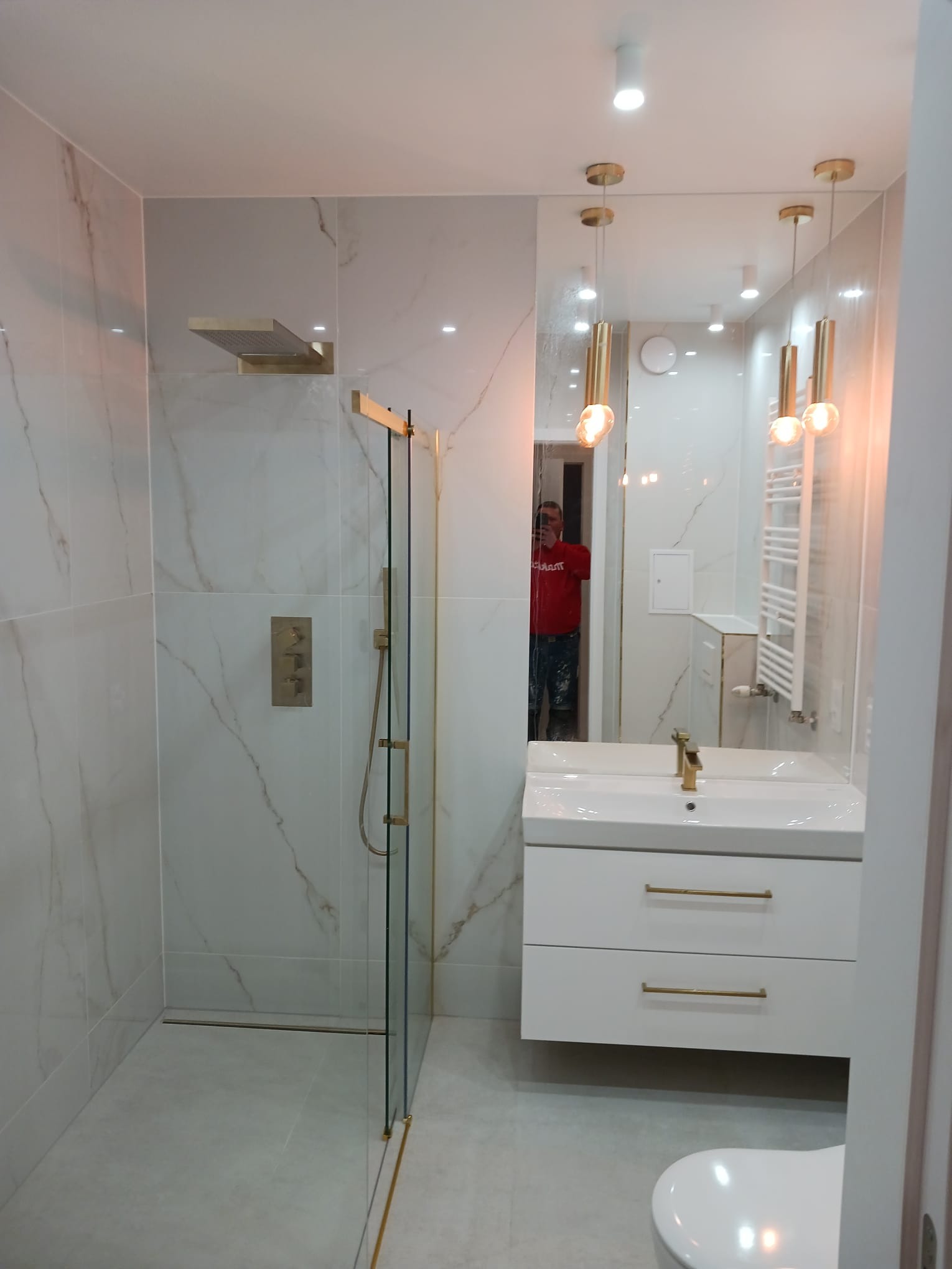 Nowoczesna łazienka z kabiną prysznicową typu walk-in, wykończona białymi płytkami imitującymi marmur z delikatnymi złotymi akcentami w armaturze i wykończeniach, widoczna umywalka z białą szafką...