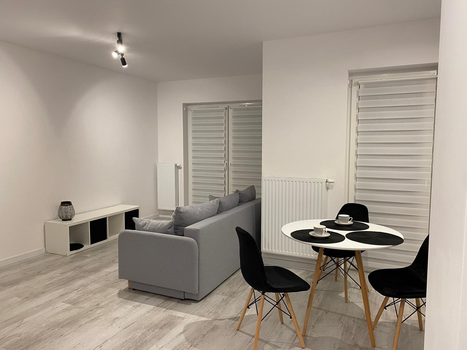 Jasne, minimalistyczne wnętrze pokoju dziennego z szarą sofą, białym stolikiem kawowym z trzema czarnymi krzesłami, białą komodą z czarnymi wnękami i drewnopodobną podłogą, oświetlone trzema...