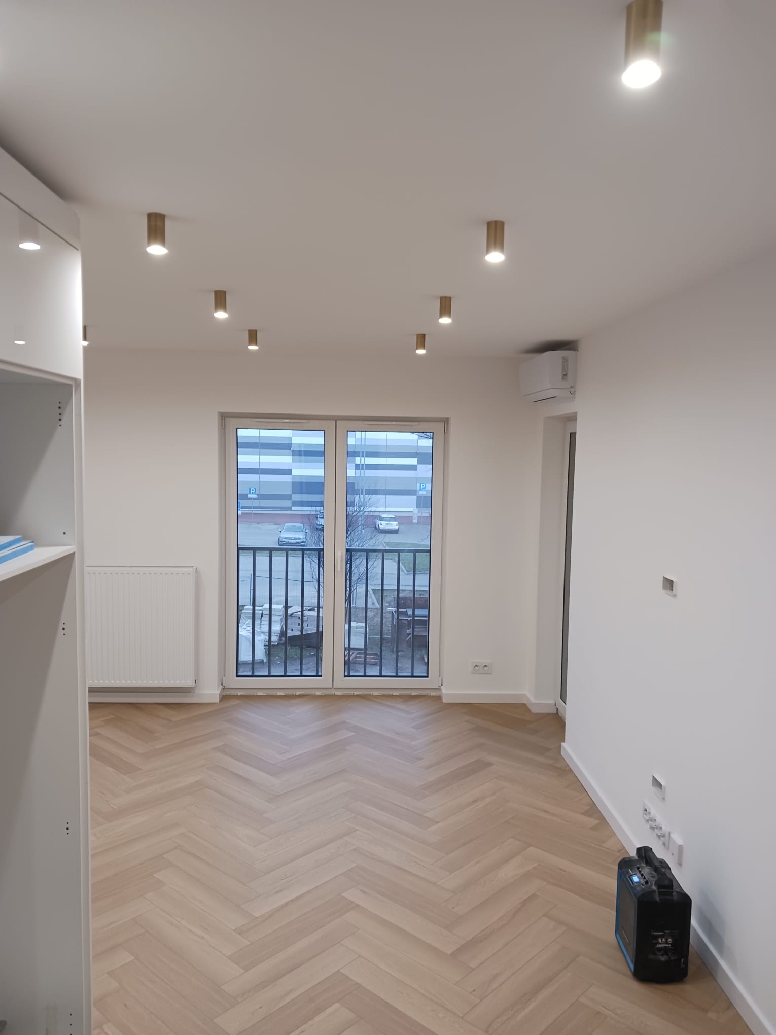 Jasne wnętrze po remoncie z parkietem ułożonym w jodełkę, białymi ścianami, oświetleniem punktowym i widokiem na parking przez balkon.
