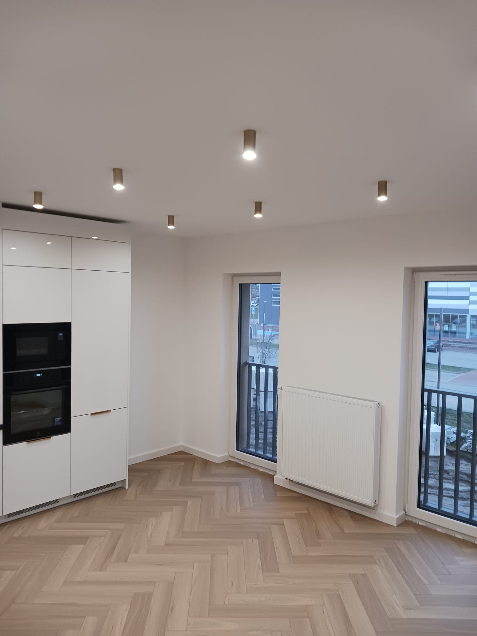 Jasne, nowocześnie wykończone wnętrze z parkietem ułożonym we wzór jodełki, białymi ścianami, zabudową kuchenną z piekarnikiem i mikrofalówką oraz dwoma oknami balkonowymi z widokiem na zewnątrz.