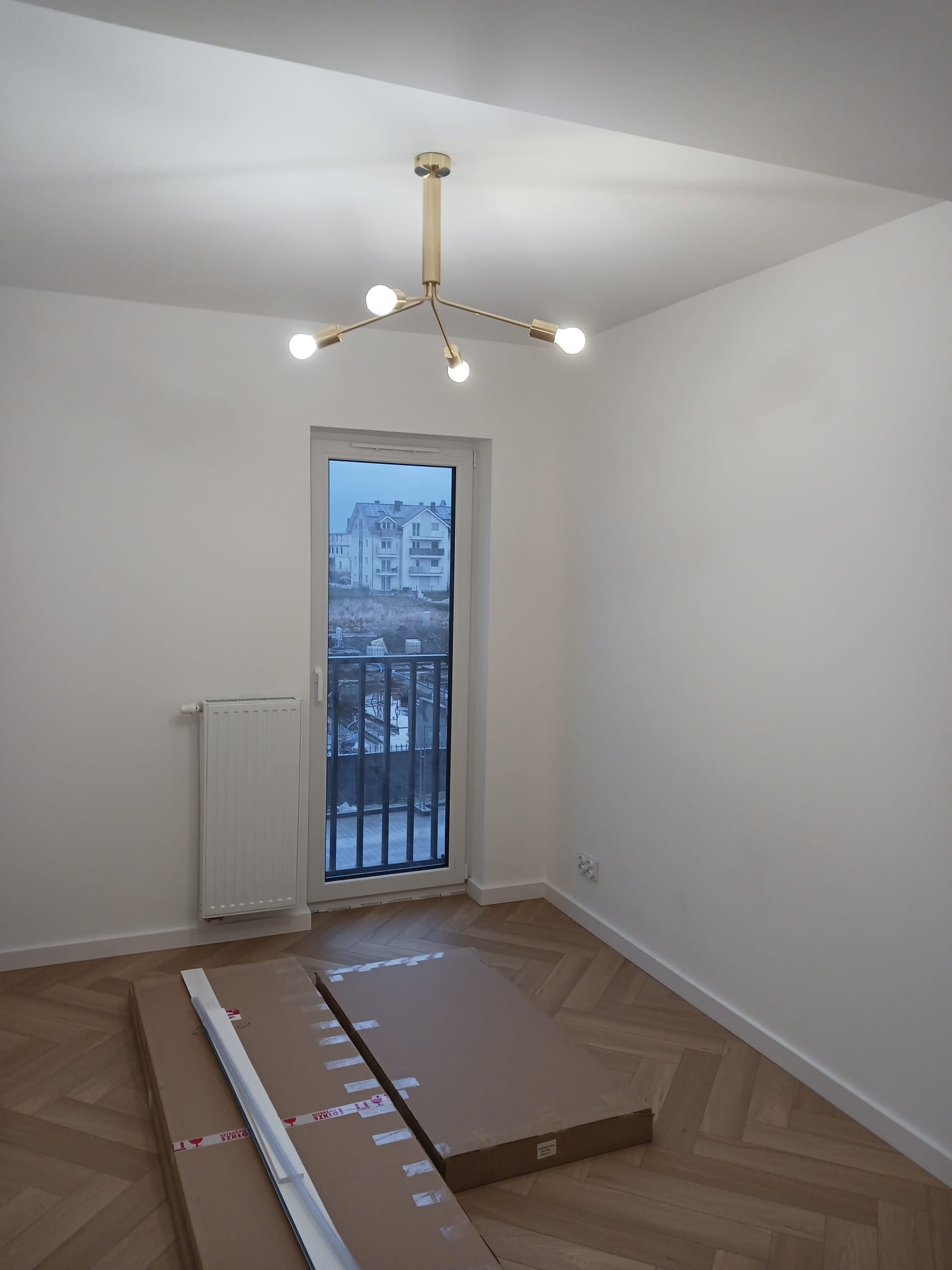 Pusty pokój z parkietem w jodełkę, świeżo pomalowanymi ścianami i nowoczesną złotą lampą sufitową. Okno z widokiem na budynek mieszkalny w tle. Na podłodze leżą paczki z materiałami wykończeniowymi.