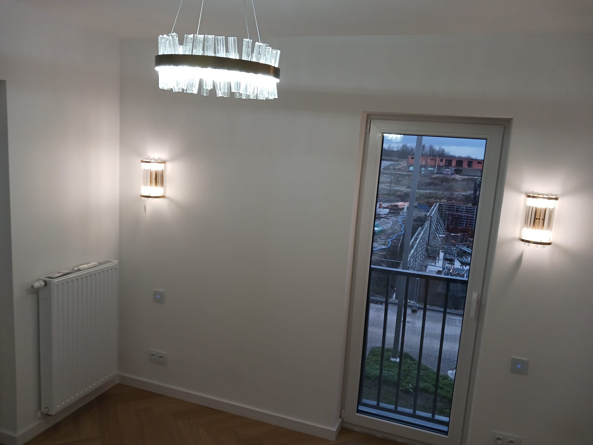 Wykończone wnętrze pokoju z parkietem w jodełkę, białymi ścianami, grzejnikiem, nowoczesnym oświetleniem sufitowym i ściennym z elementami szkła, oraz oknem balkonowym z widokiem na teren budowy.