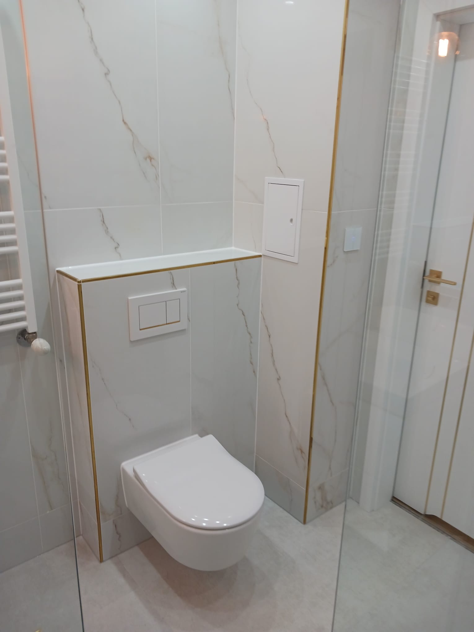 Nowoczesna toaleta podwieszana w łazience wykończonej białymi płytkami imitującymi marmur z delikatnymi złotymi akcentami, widoczny grzejnik drabinkowy i szklana kabina prysznicowa.