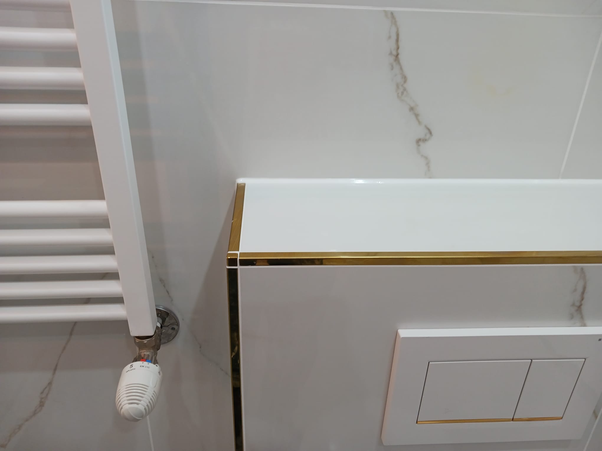 Nowoczesna łazienka z białymi płytkami imitującymi marmur, grzejnikiem drabinkowym i zabudową WC z akcentami w kolorze złota.