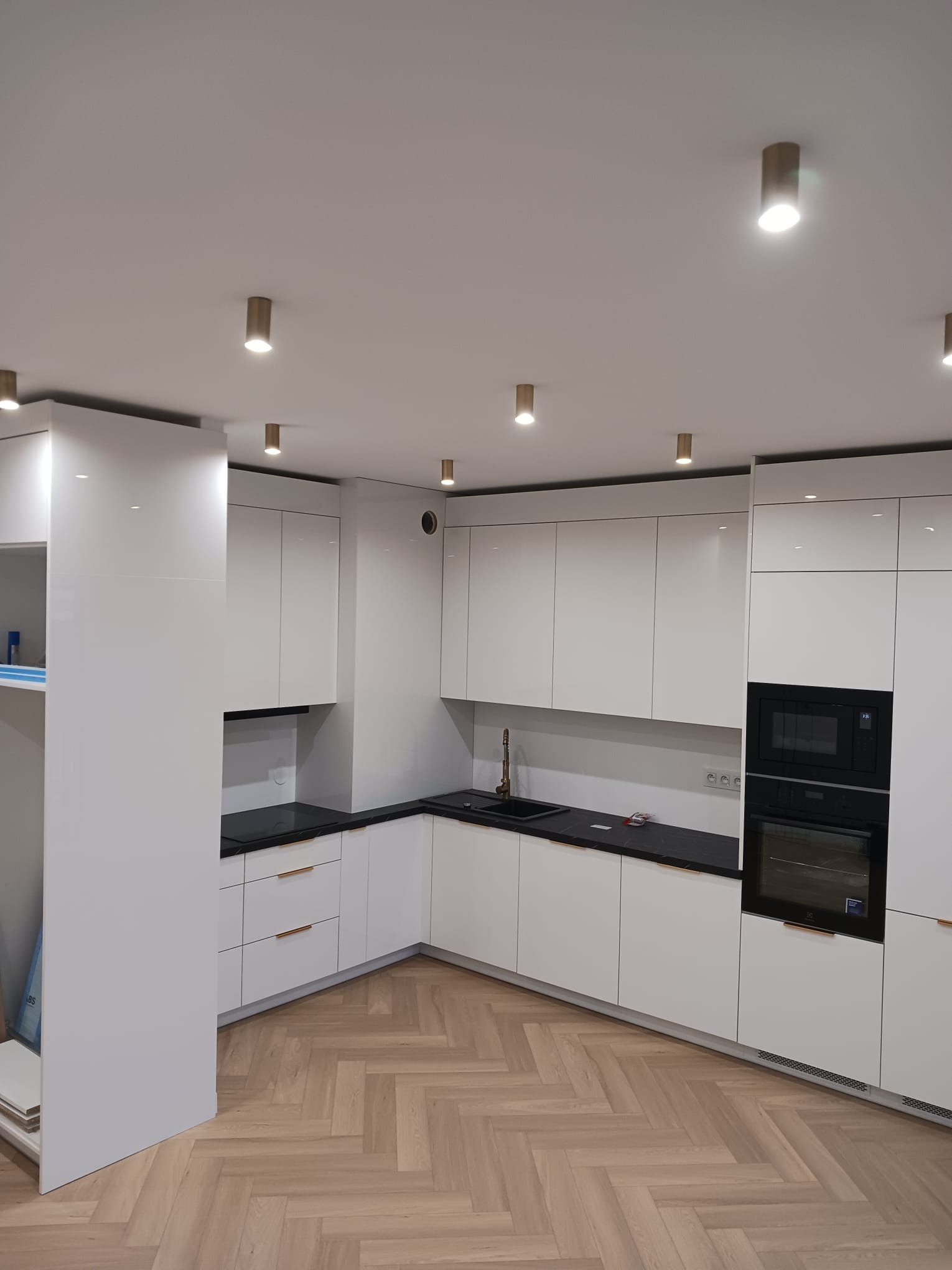 Nowoczesna, biała kuchnia z czarnymi blatami i parkietem ułożonym w jodełkę. Złote detale, takie jak uchwyty i bateria, dodają elegancji. Punktowe oświetlenie sufitowe. Widoczny piekarnik i płyta...