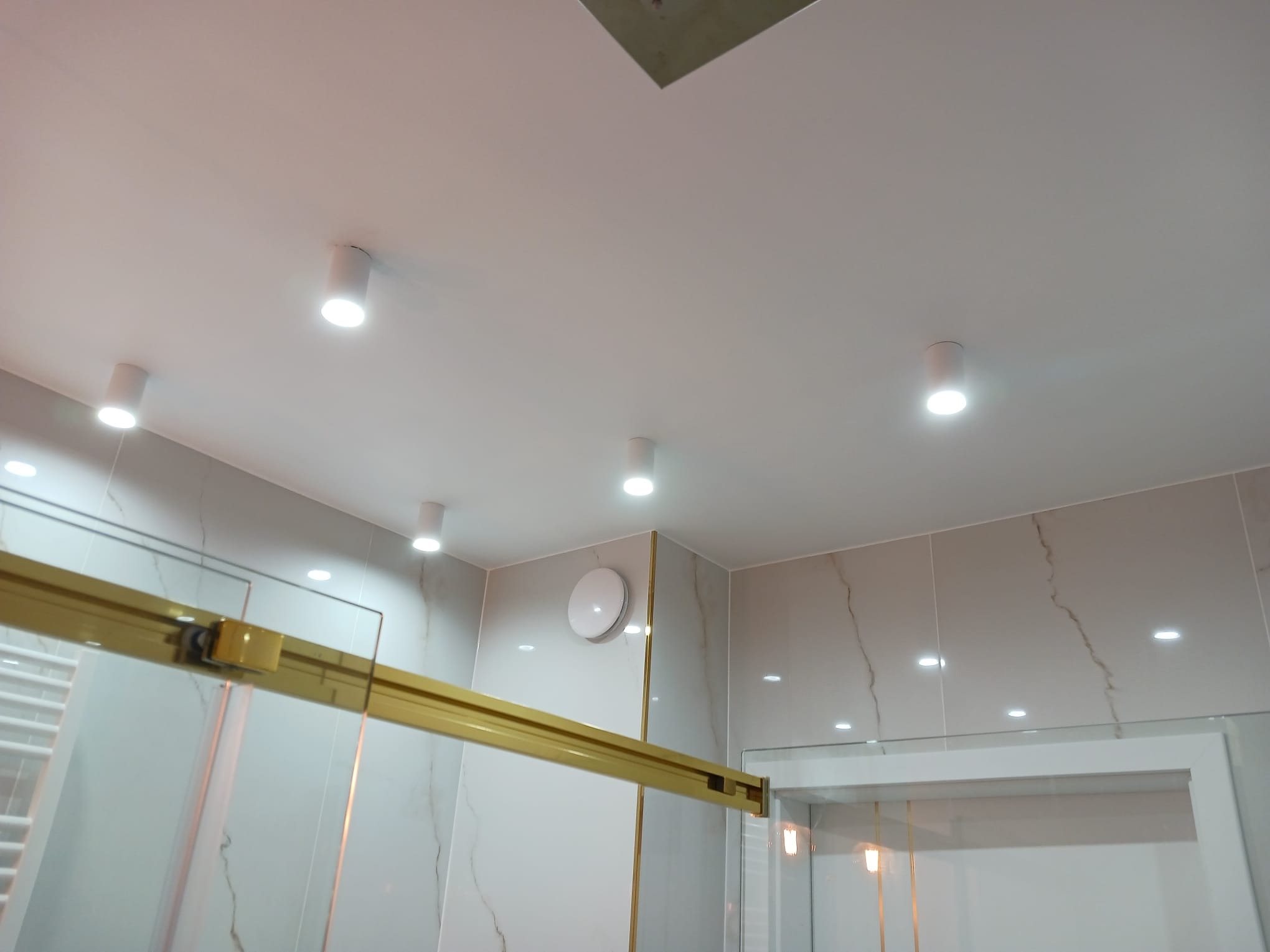 Wykończona łazienka z jasnymi płytkami imitującymi marmur, złotymi akcentami na kabinie prysznicowej i punktowym oświetleniem sufitowym.