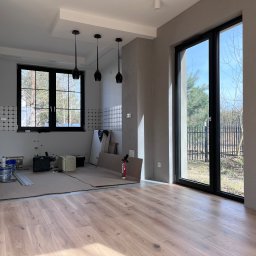 Just_Bud - Wnętrze w trakcie remontu: położona drewniana podłoga, zabezpieczona podłoga w części kuchennej, widoczne narzędzia i materiały budowlane, surowe ściany w odcieniach szarości z nowoczesnym...