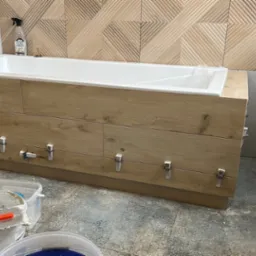 Montaż płytek imitujących drewno na obudowie wanny, widoczne klipsy poziomujące, w tle dekoracyjne płytki ścienne, Żabia Wola.