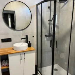 Nowoczesna łazienka z kabiną prysznicową z czarnymi profilami, okrągłym lustrem i białą umywalką nablatową na szafce z drewnianym blatem, wykończona szarą płytką.