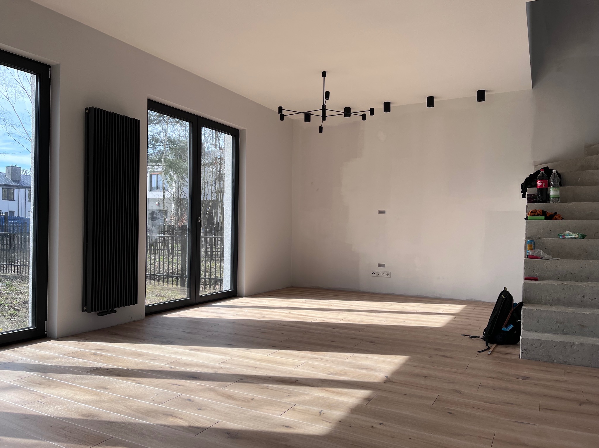 Jasne, przestronne wnętrze z drewnopodobną podłogą, dużymi oknami z czarnymi ramami, czarnym grzejnikiem i nowoczesną lampą sufitową. Schody prowadzące na górę w stanie surowym z widocznymi...