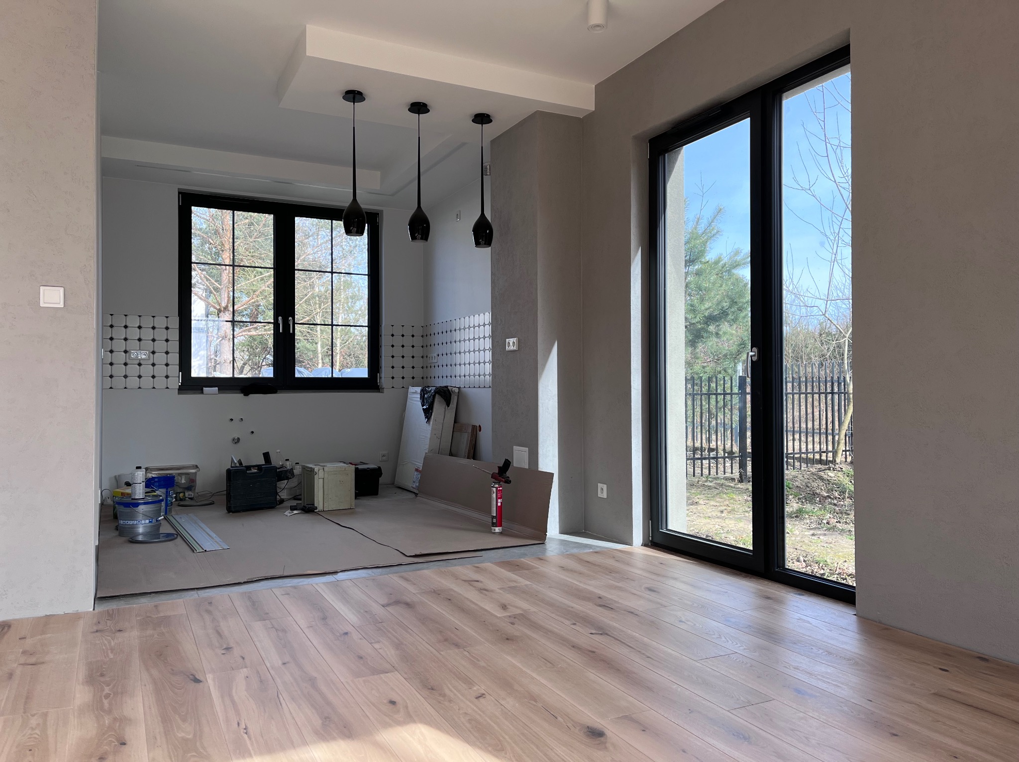 Wnętrze w trakcie remontu: położona drewniana podłoga, zabezpieczona podłoga w części kuchennej, widoczne narzędzia i materiały budowlane, surowe ściany w odcieniach szarości z nowoczesnym...