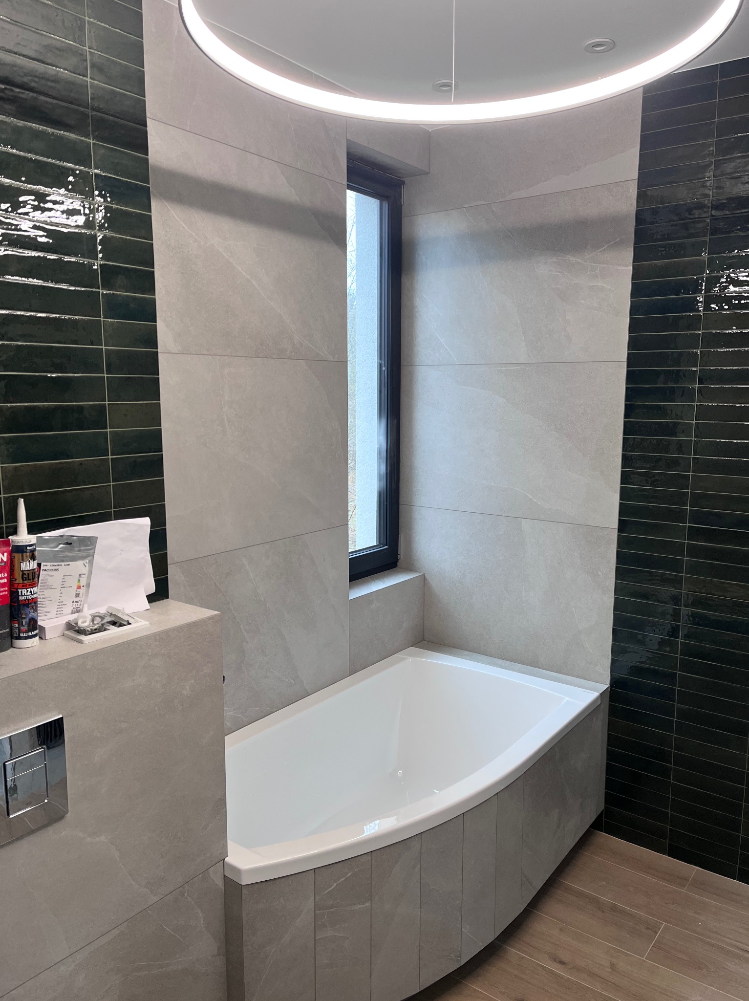 Nowoczesna łazienka z wanną obłożoną płytkami imitującymi marmur, kontrastującą ścianą z ciemnozielonych, błyszczących płytek ułożonych poziomo, oraz okrągłym, podwieszanym oświetleniem LED.