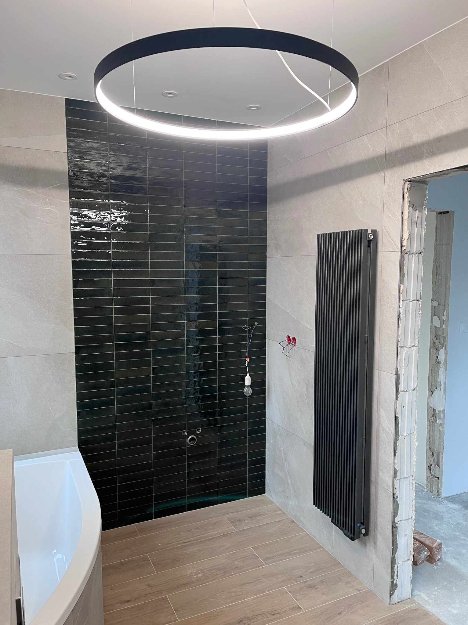 Nowoczesna łazienka w trakcie aranżacji: czarne, błyszczące płytki na ścianie, biała wanna, pionowy czarny grzejnik, okrągła lampa sufitowa i drewnopodobne płytki na podłodze, widoczne przewody...