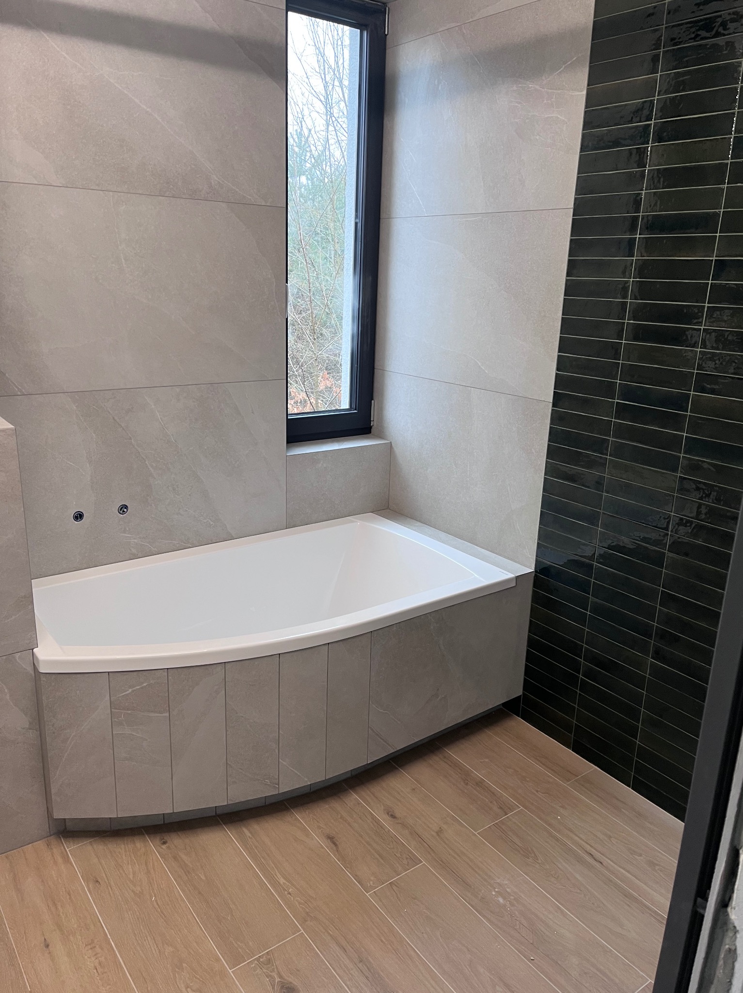 Nowoczesna łazienka z wanną obudowaną płytkami imitującymi kamień, jasnymi płytkami na ścianach, ciemnymi, wąskimi płytkami w pionowym pasie oraz drewnopodobną podłogą. Okno z widokiem na drzewa.