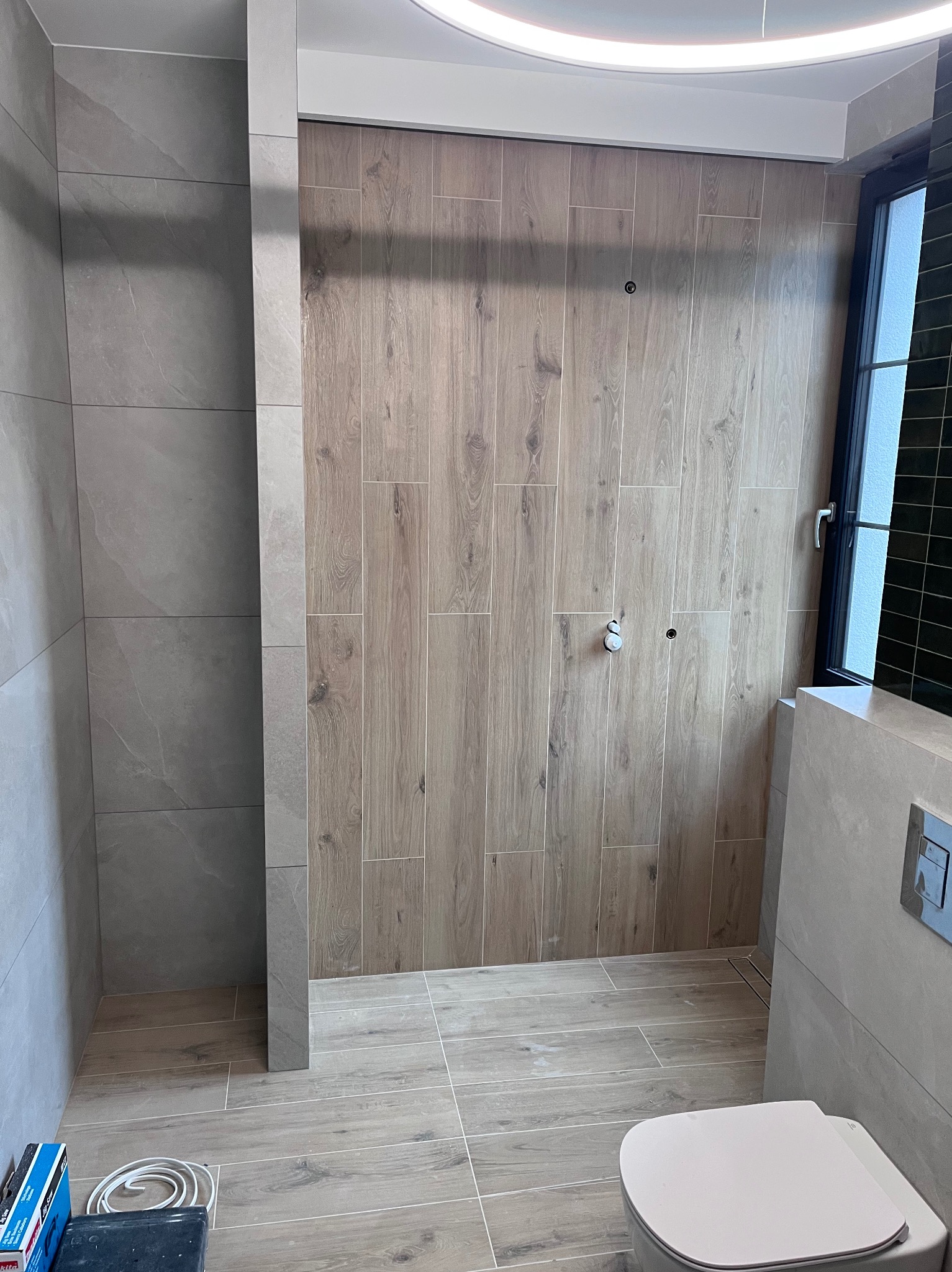 Łazienka w trakcie wykańczania: drewnopodobne płytki na ścianach i podłodze, widoczne przyłącza hydrauliczne, częściowo zamontowana toaleta podwieszana, czarne płytki w obramowaniu okna, okrągła...