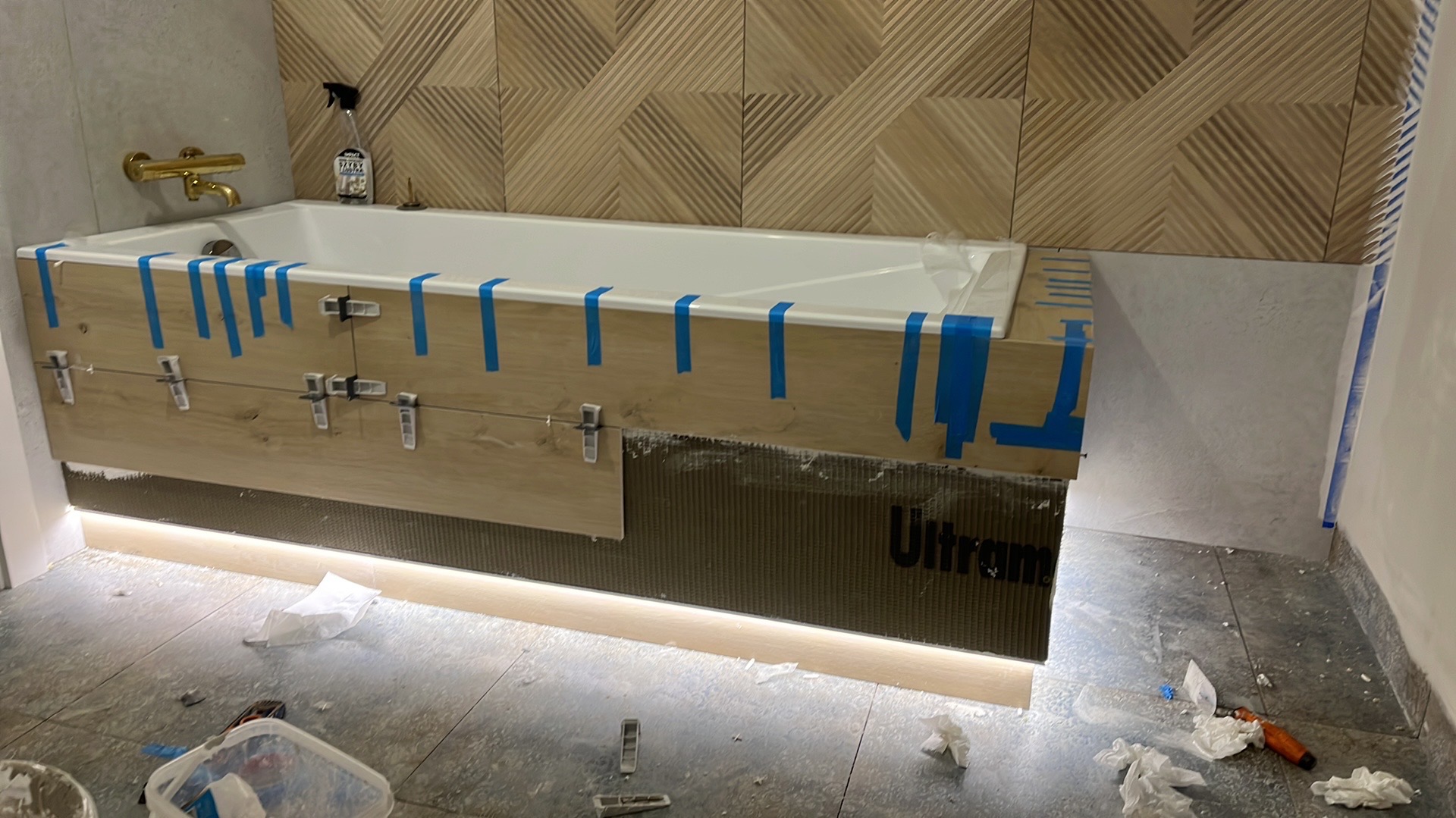 Montaż płytek imitujących drewno na obudowie wanny z podświetleniem LED, widoczne klipsy poziomujące, taśma malarska i narzędzia na podłodze.