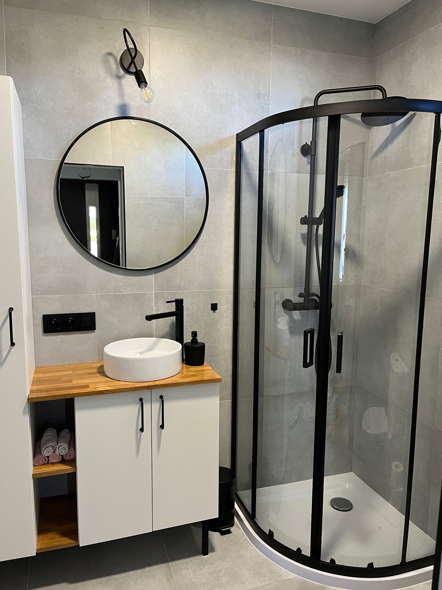 Nowoczesna łazienka z szarą płytką, okrągłym lustrem w czarnej ramie, białą umywalką nablatową na drewnianym blacie, czarną armaturą i kabiną prysznicową z czarnymi profilami.