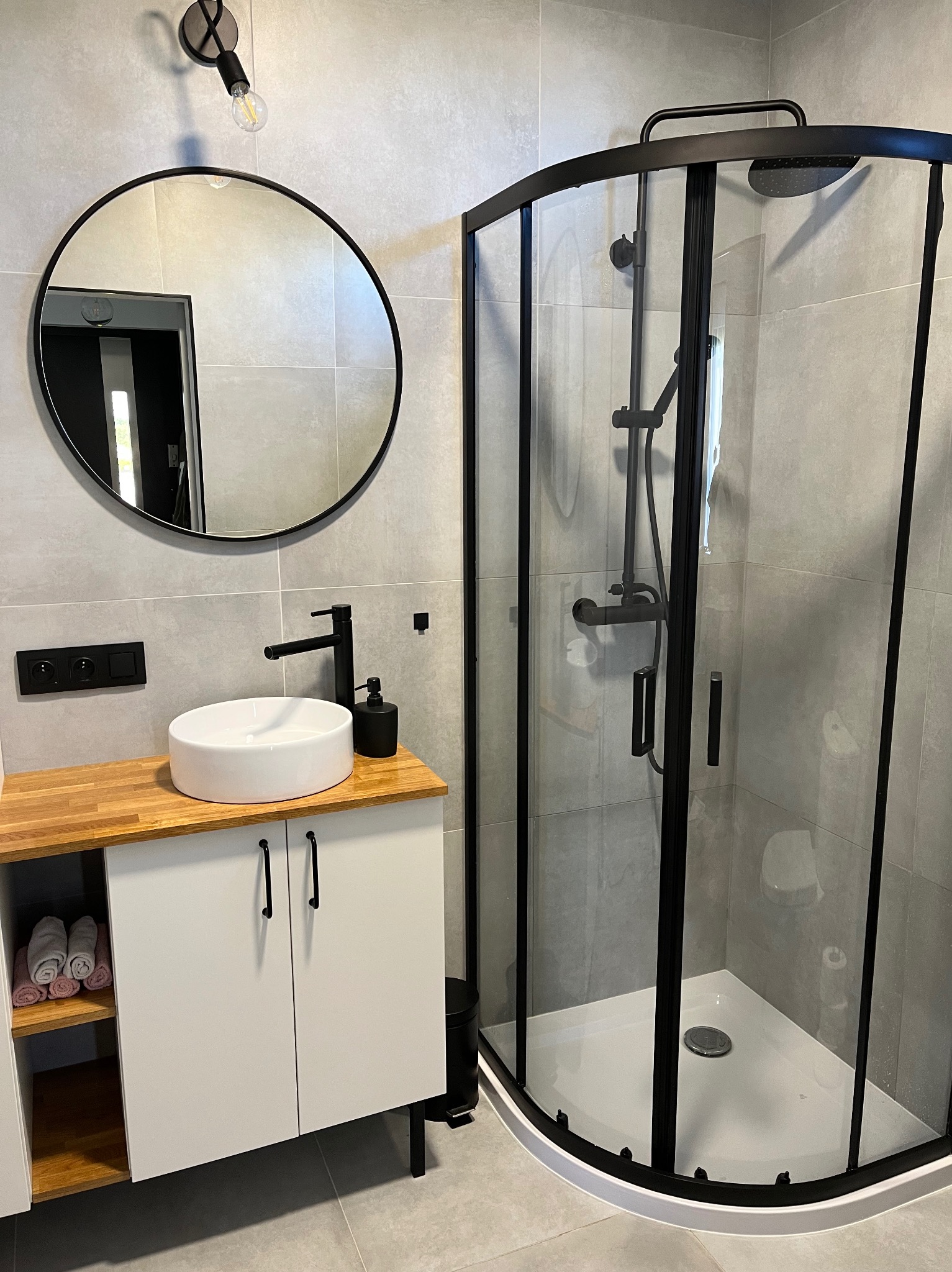 Nowoczesna łazienka z kabiną prysznicową z czarnymi profilami, okrągłym lustrem i białą umywalką nablatową na szafce z drewnianym blatem, wykończona szarą płytką.