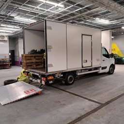 Qpack Diana Waliwender - Biały samochód dostawczy z otwartymi drzwiami załadunkowymi i rampą, częściowo załadowany paletami i workami, w hali garażowej z kratownicą sufitową.