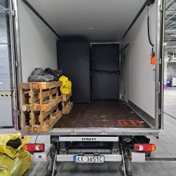 Qpack Diana Waliwender - Wnętrze paki busa transportowego z otwartymi drzwiami, widoczne palety drewniane z kocami i torbami, czarne szafy w środku, podnośnik hydrauliczny na zewnątrz i tablica rejestracyjna.