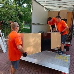 Qpack Diana Waliwender - Trzech mężczyzn w pomarańczowych koszulkach przenosi duży, płaski element meblowy z ciężarówki na rampę, widoczne drzewa w tle.