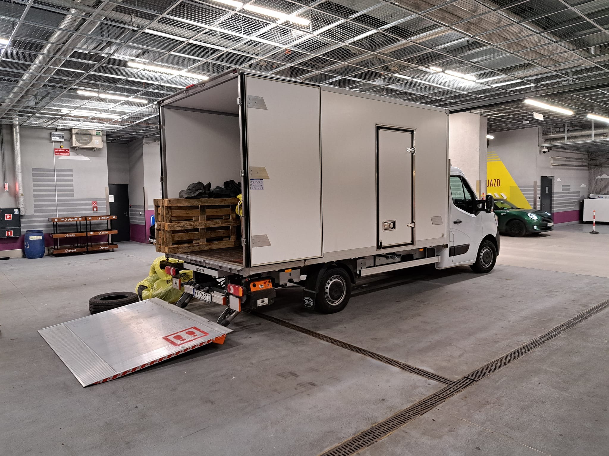Biały samochód dostawczy z otwartymi drzwiami załadunkowymi i rampą, częściowo załadowany paletami i workami, w hali garażowej z kratownicą sufitową.