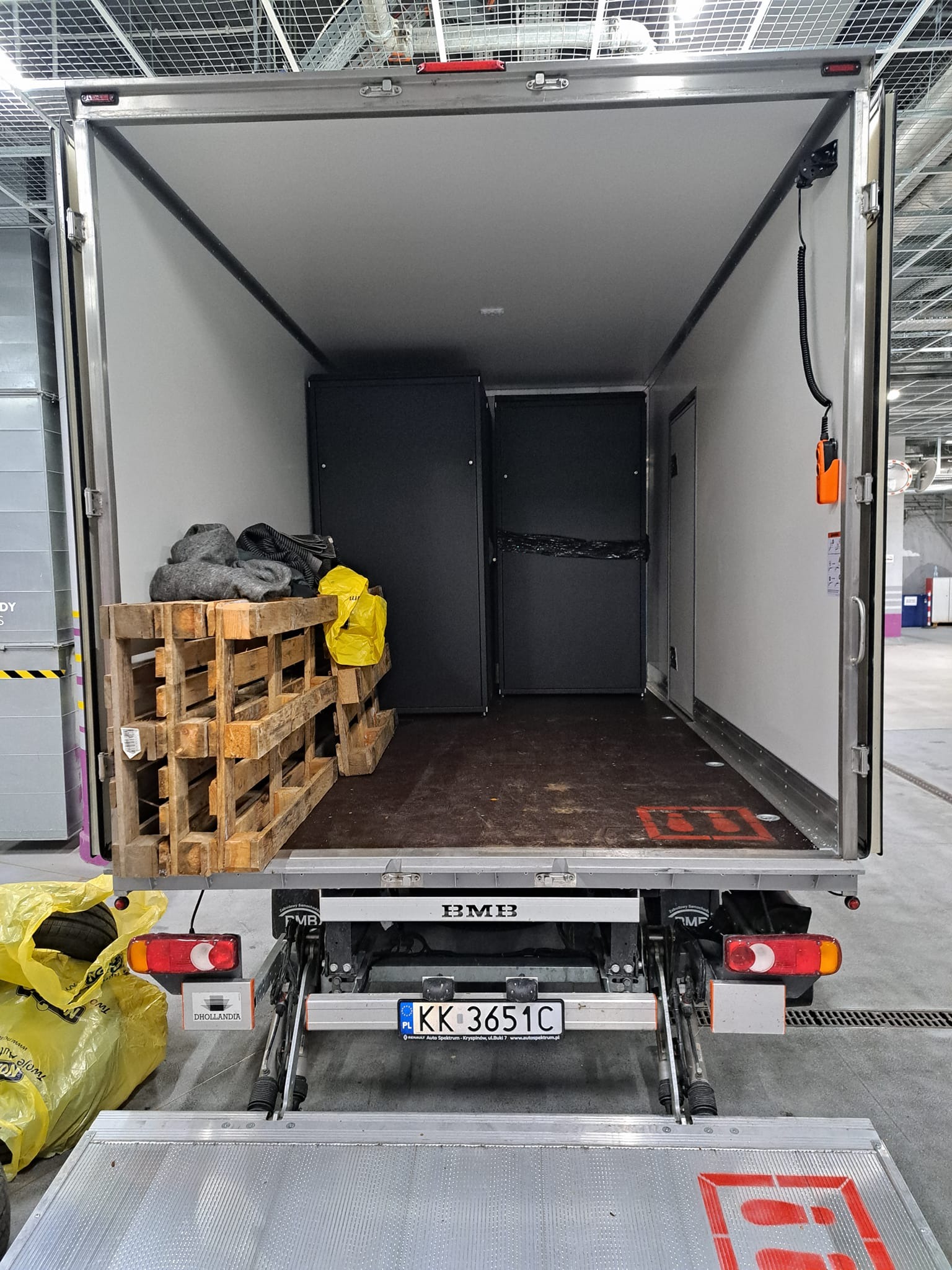 Wnętrze paki busa transportowego z otwartymi drzwiami, widoczne palety drewniane z kocami i torbami, czarne szafy w środku, podnośnik hydrauliczny na zewnątrz i tablica rejestracyjna.