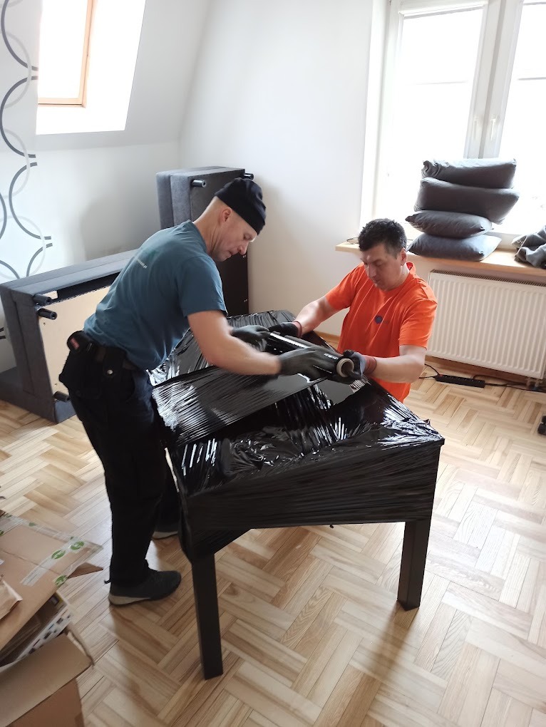 Dwóch mężczyzn w rękawicach owija czarną folią stretch stół przed transportem w jasnym pomieszczeniu z parkietem.