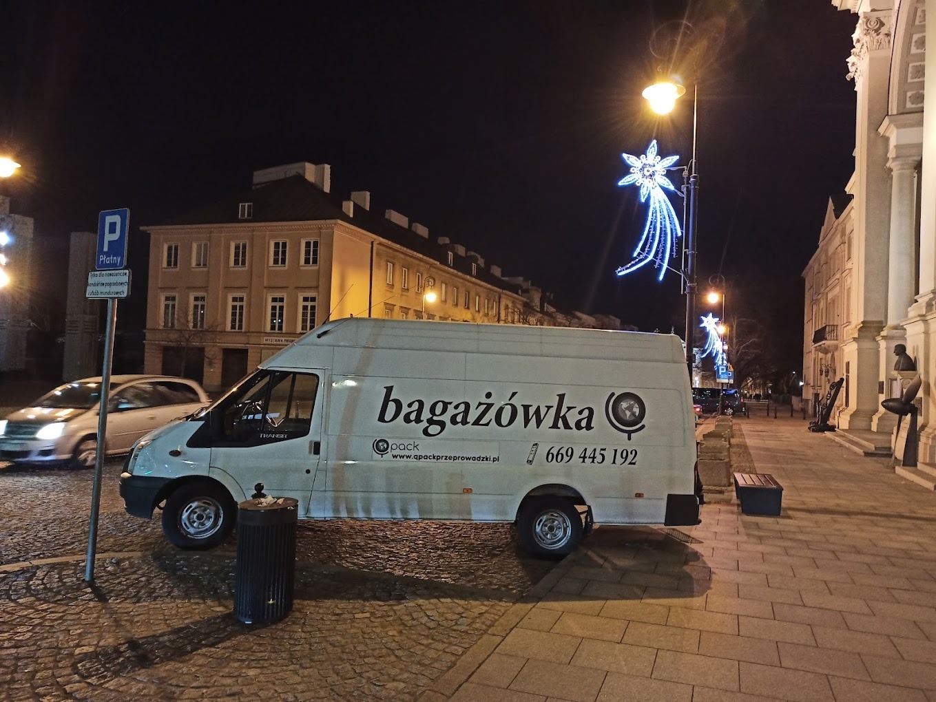 Biały bus z napisem 'bagażówka' i numerem telefonu zaparkowany na brukowanej ulicy w mieście nocą, widoczne dekoracje świąteczne na latarniach.