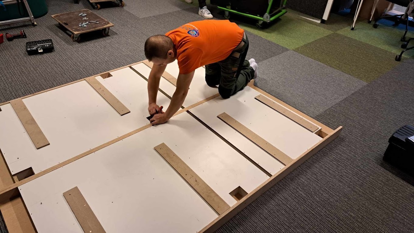 Pracownik w pomarańczowej koszulce montuje elementy meblowe na podłodze w biurze, przygotowując je do transportu.