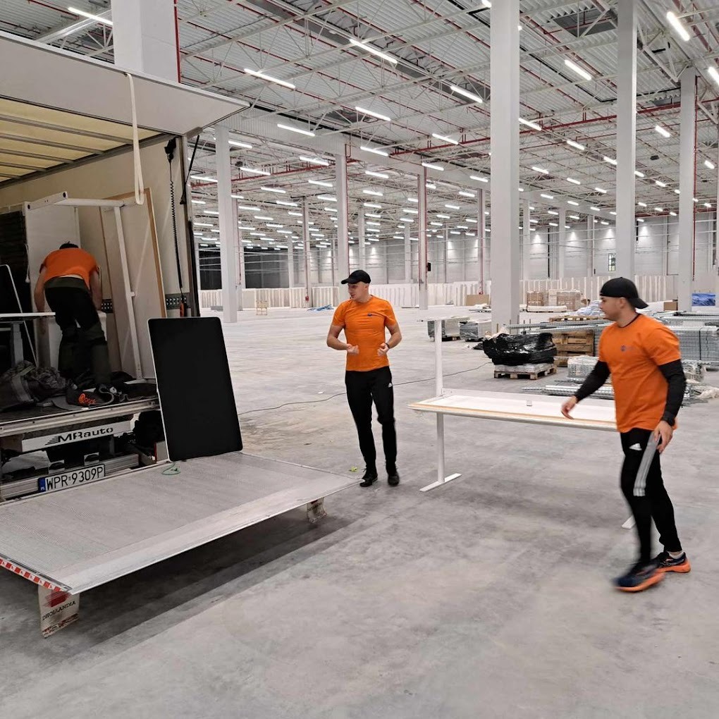 Pracownicy w pomarańczowych koszulkach rozładowują biurka z przyczepy wewnątrz hali magazynowej z widoczną konstrukcją dachu i kolumnami.